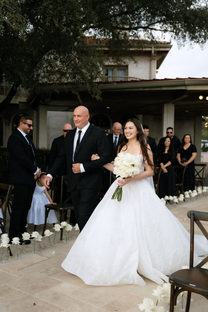 Bride in white gown standing against stone arches at Villa Antonia with black-dressed wedding party