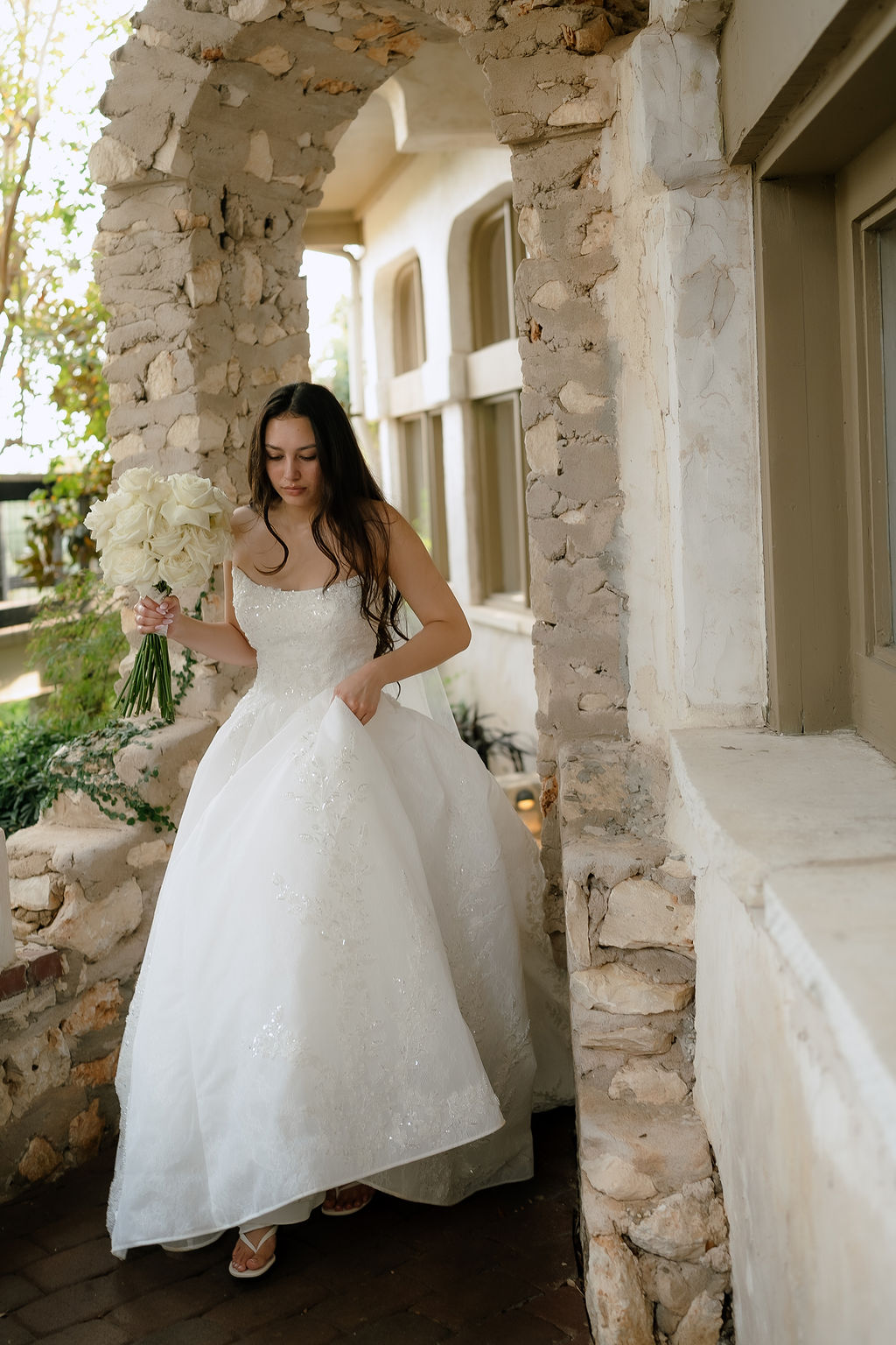 Bride in white gown standing against stone arches at Villa Antonia with black-dressed wedding party