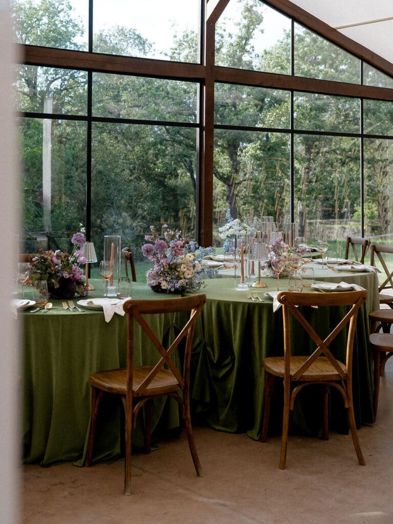 Wide angle view of Monet inspired wedding editorial tablescape at Wild Roots Lockhart Texas featuring sage green linens, garden floral centerpieces, and floor-to-ceiling windows overlooking wooded landscape, photographed by Austin wedding photographer Andrea Jang