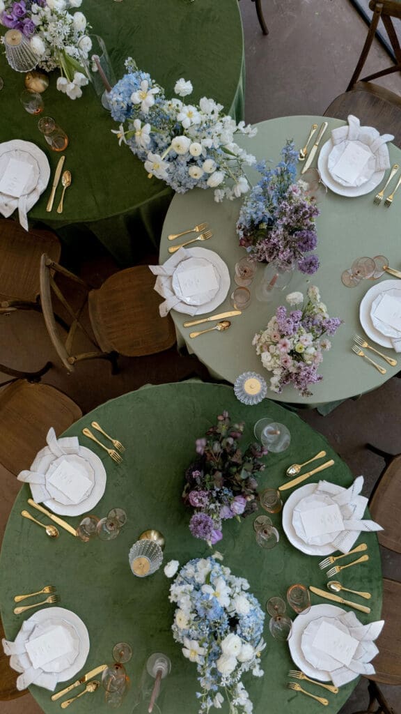 Wide angle view of Monet inspired wedding editorial tablescape at Wild Roots Lockhart Texas featuring sage green linens, garden floral centerpieces, and floor-to-ceiling windows overlooking wooded landscape, photographed by Austin wedding photographer Andrea Jang