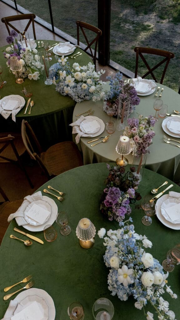 Wide angle view of Monet inspired wedding editorial tablescape at Wild Roots Lockhart Texas featuring sage green linens, garden floral centerpieces, and floor-to-ceiling windows overlooking wooded landscape, photographed by Austin wedding photographer Andrea Jang